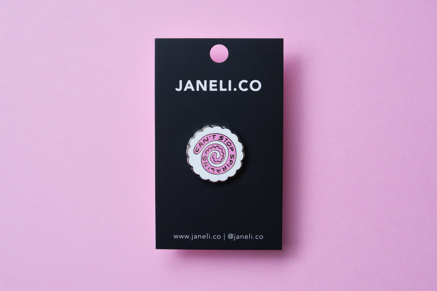 An enamel pin showing a narutomaki fishcake that says "Can't Stop Spiraling" on a black JaneLi.Co backing card over a pink background.