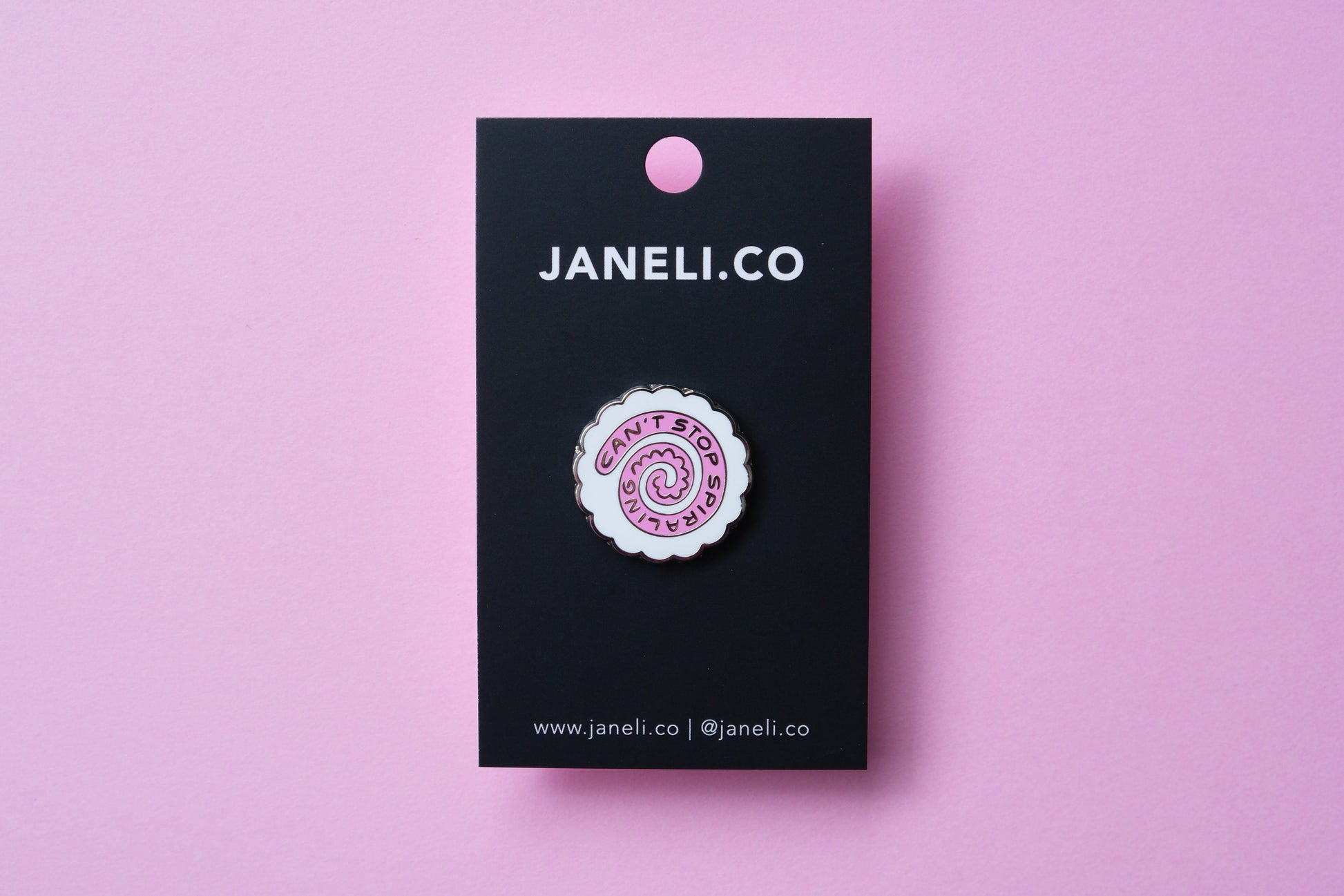 An enamel pin showing a narutomaki fishcake that says "Can't Stop Spiraling" on a black JaneLi.Co backing card over a pink background.