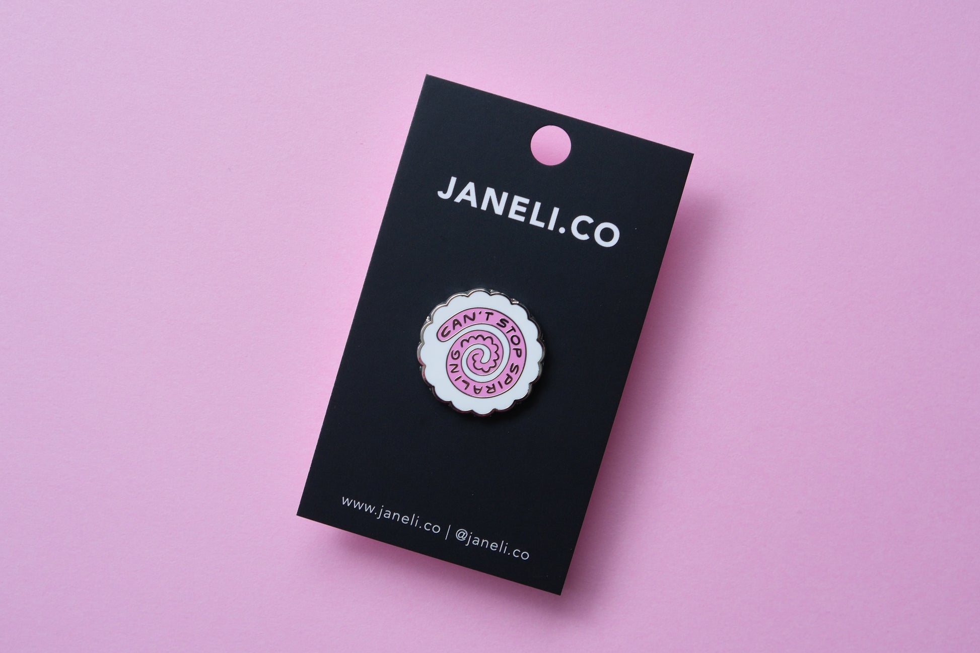 An enamel pin showing a narutomaki fishcake that says "Can't Stop Spiraling" on a black JaneLi.Co backing card over a pink background.