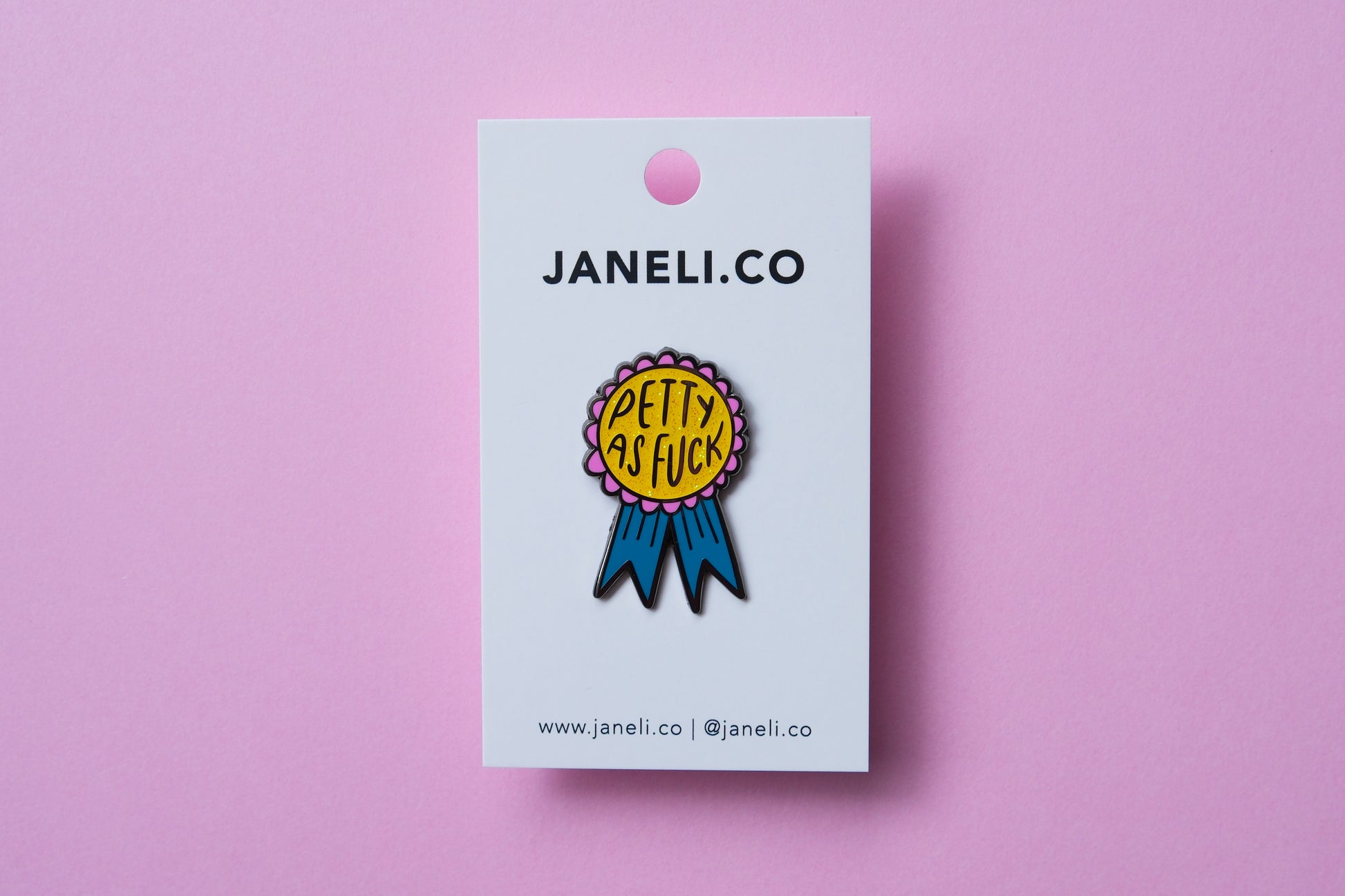 An enamel pin showing an award ribbon that says "Petty As Fuck" on a white JaneLi.Co backing card over a pink background.