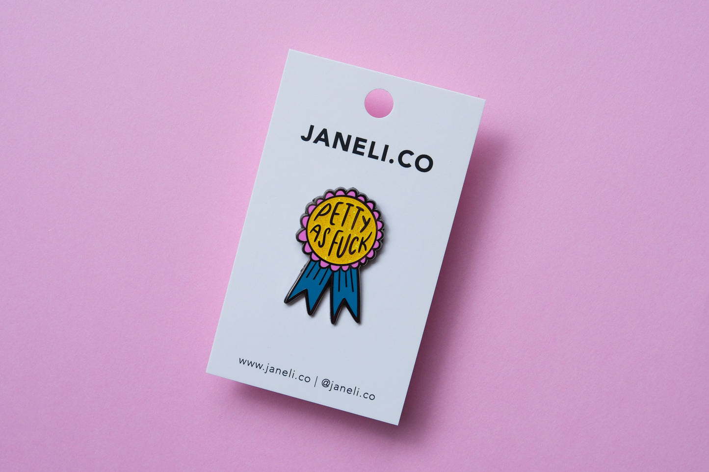 An enamel pin showing an award ribbon that says "Petty As Fuck" on a white JaneLi.Co backing card over a pink background.