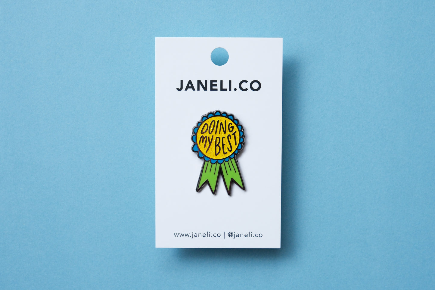 An enamel pin showing an award ribbon that says "Doing My Best" on a white JaneLi.Co backing card over a blue background.