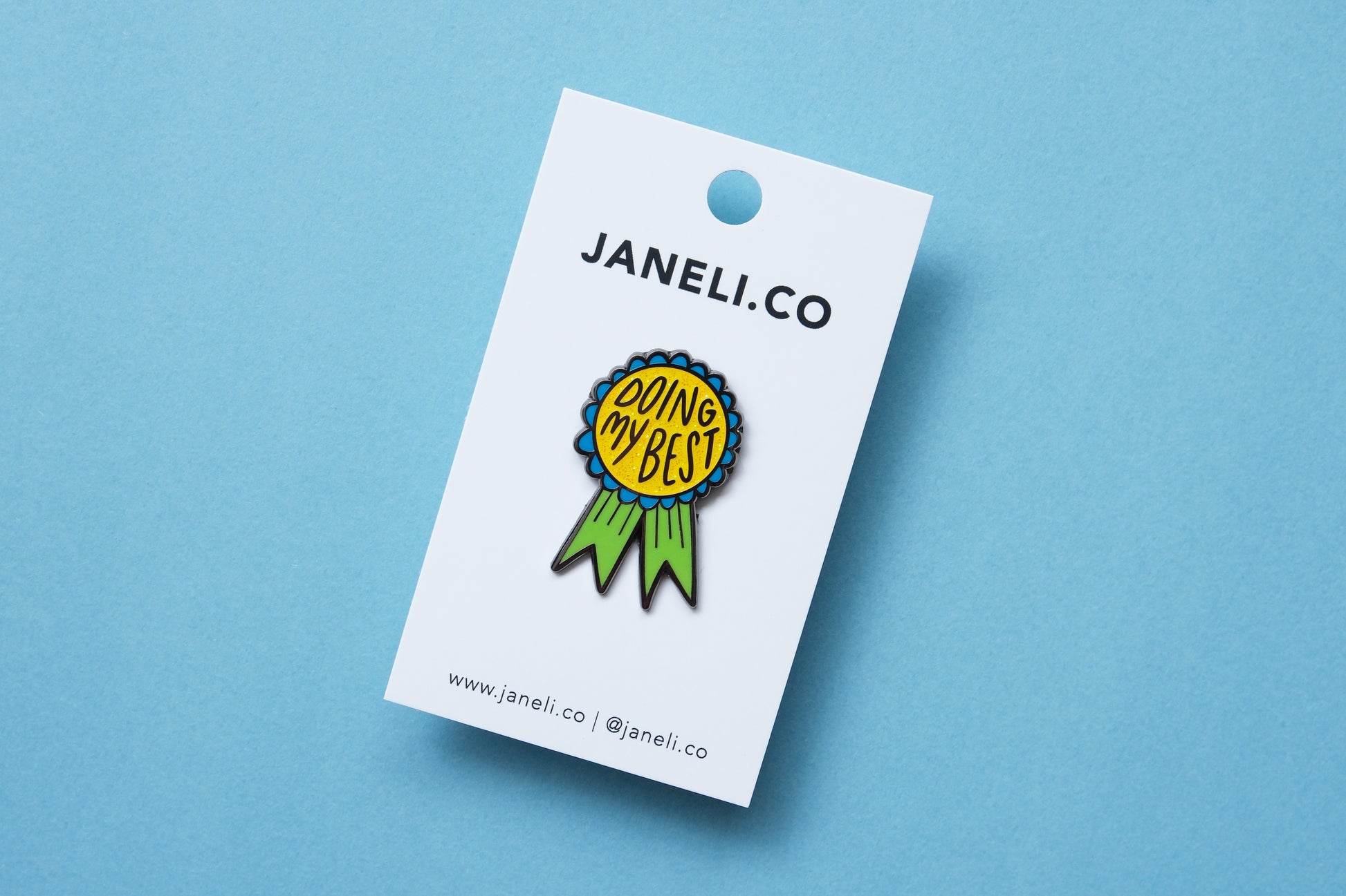 An enamel pin showing an award ribbon that says "Doing My Best" on a white JaneLi.Co backing card over a blue background.