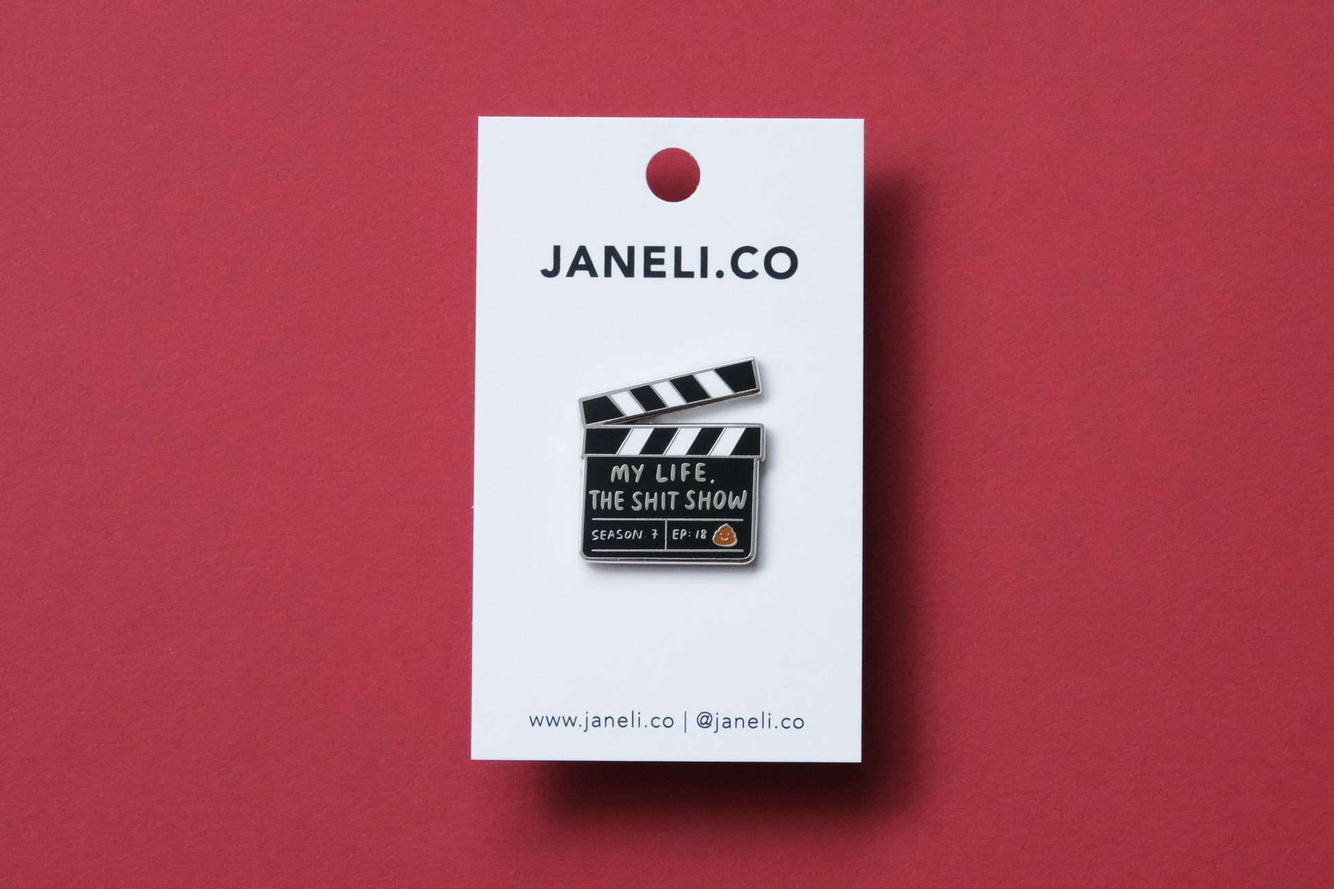 An enamel pin showing a movie clapper that says "My Life, the Shit Show" on a white JaneLi.Co backing card over a maroon background.