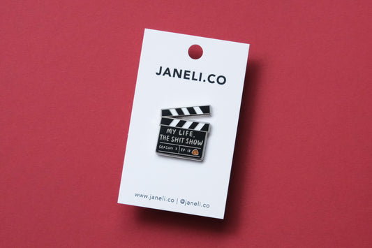 An enamel pin showing a movie clapper that says "My Life, the Shit Show" on a white JaneLi.Co backing card over a maroon background.