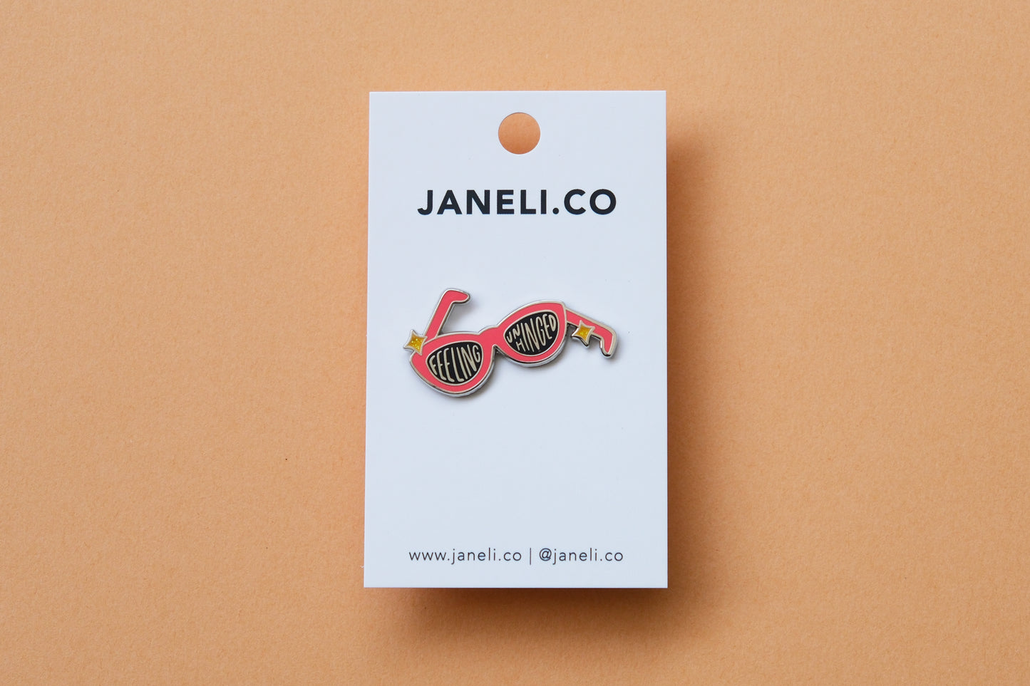 An enamel pin showing a pair of broken sunglasses that say "Feeling Unhinged" on a white JaneLi.Co backing card over a orange background.