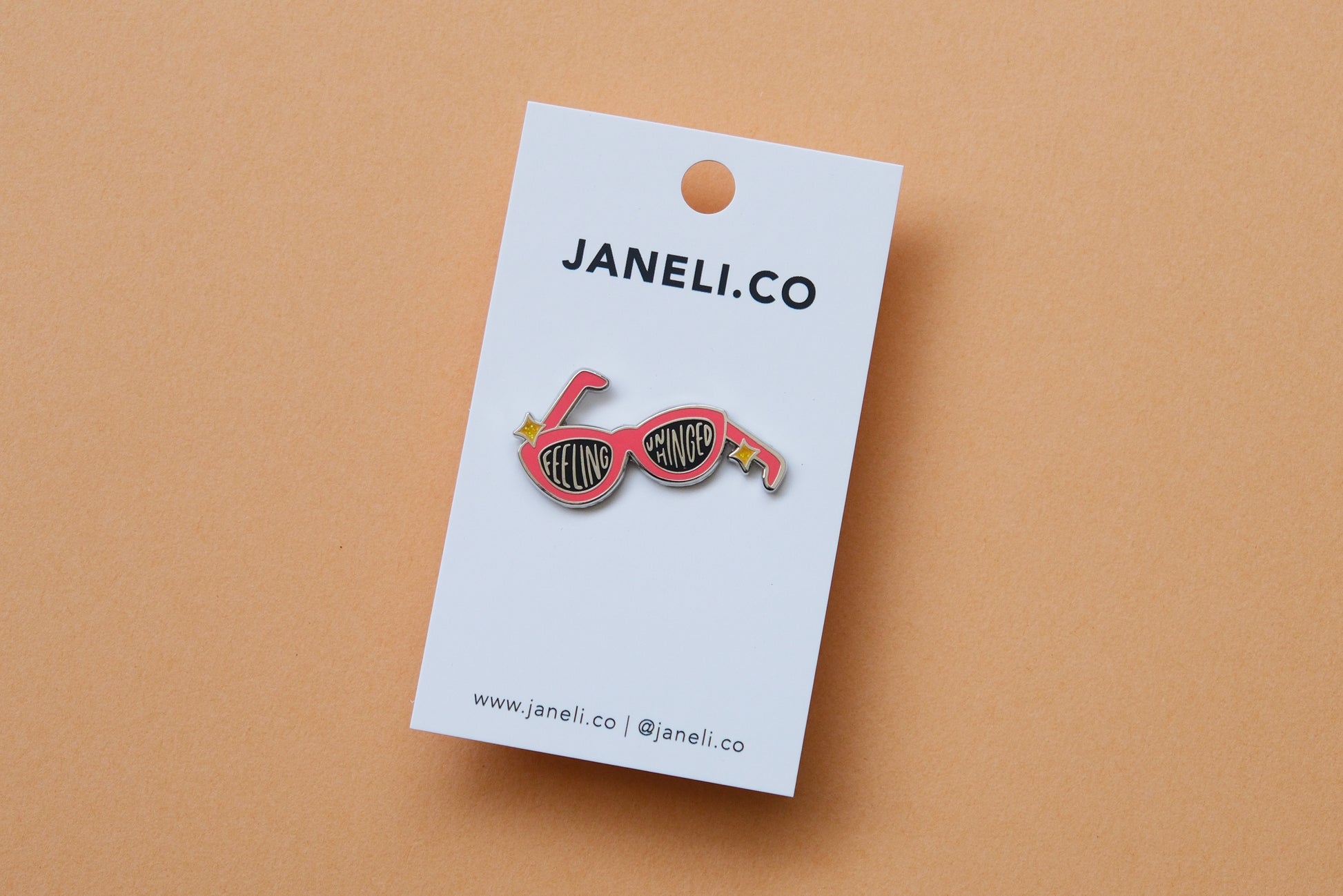 An enamel pin showing a pair of broken sunglasses that say "Feeling Unhinged" on a white JaneLi.Co backing card over a orange background.