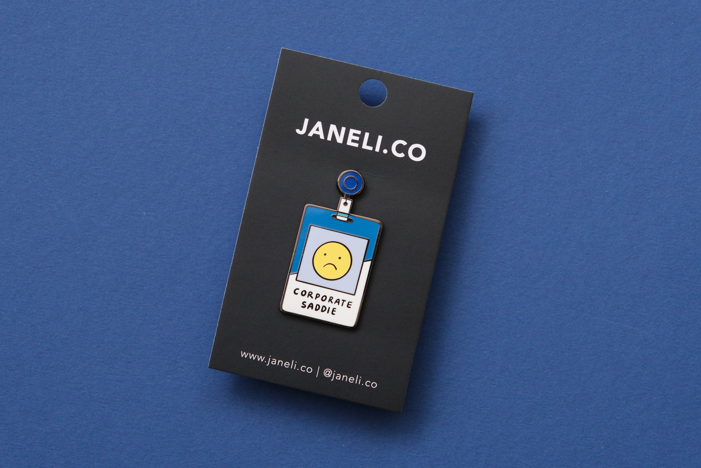 An enamel pin showing a corporate badge that says "Corporate Saddie" with a sad face on a black JaneLi.Co backing card over a blue background.