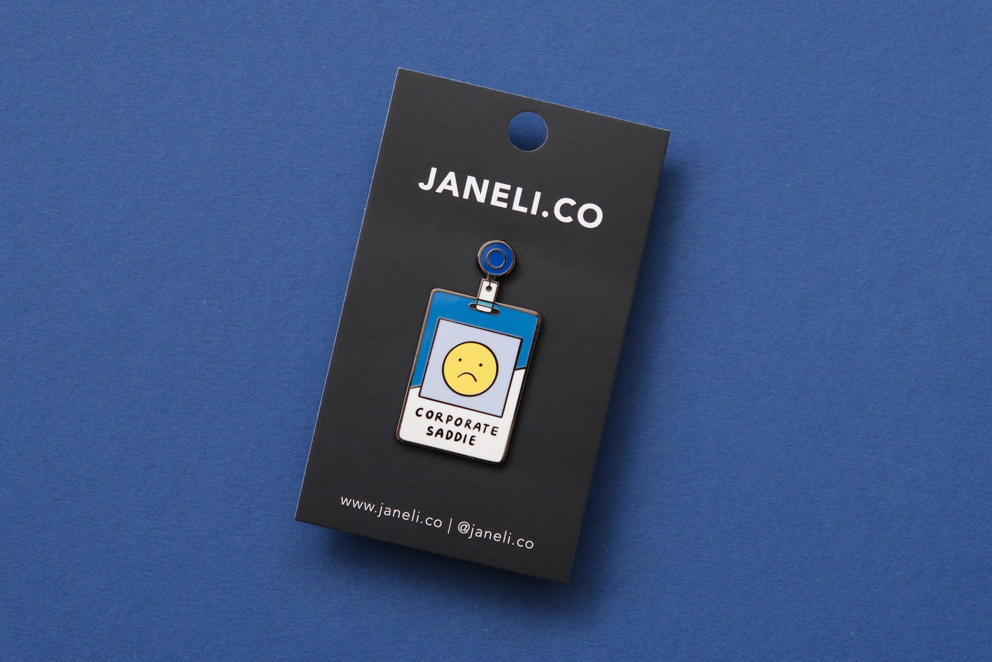 An enamel pin showing a corporate badge that says "Corporate Saddie" with a sad face on a black JaneLi.Co backing card over a blue background.