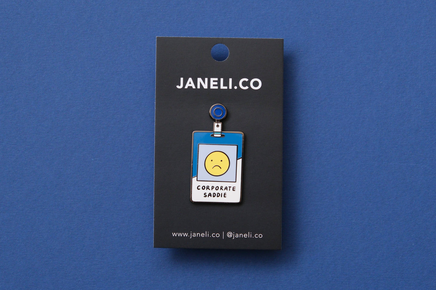 An enamel pin showing a corporate badge that says "Corporate Saddie" with a sad face on a black JaneLi.Co backing card over a blue background.