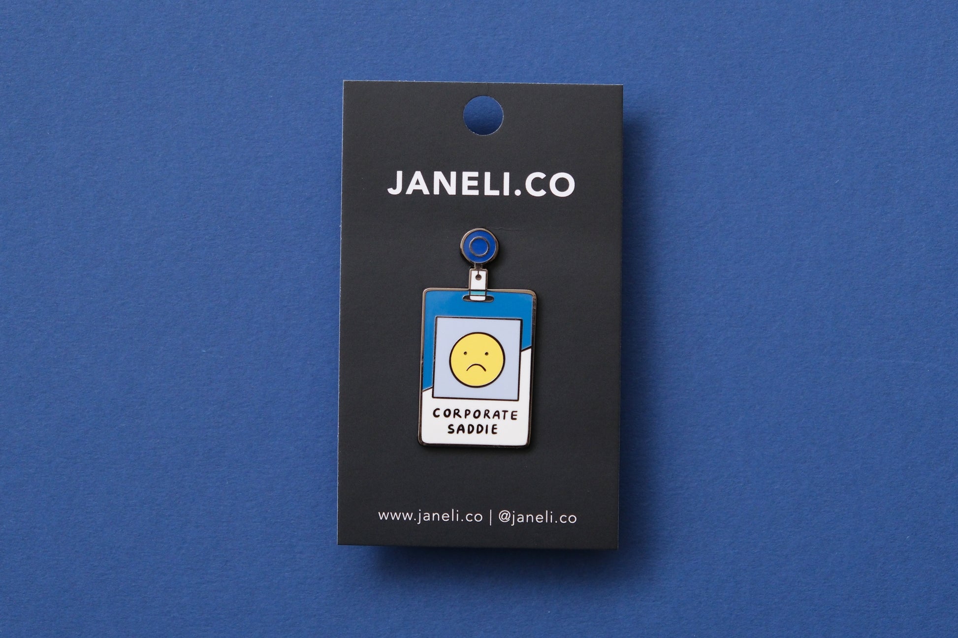 An enamel pin showing a corporate badge that says "Corporate Saddie" with a sad face on a black JaneLi.Co backing card over a blue background.