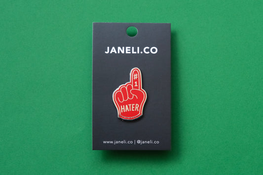 An enamel pin showing a rerd foam finger that says "#1 Hater" on a black JaneLi.Co backing card over a green background.