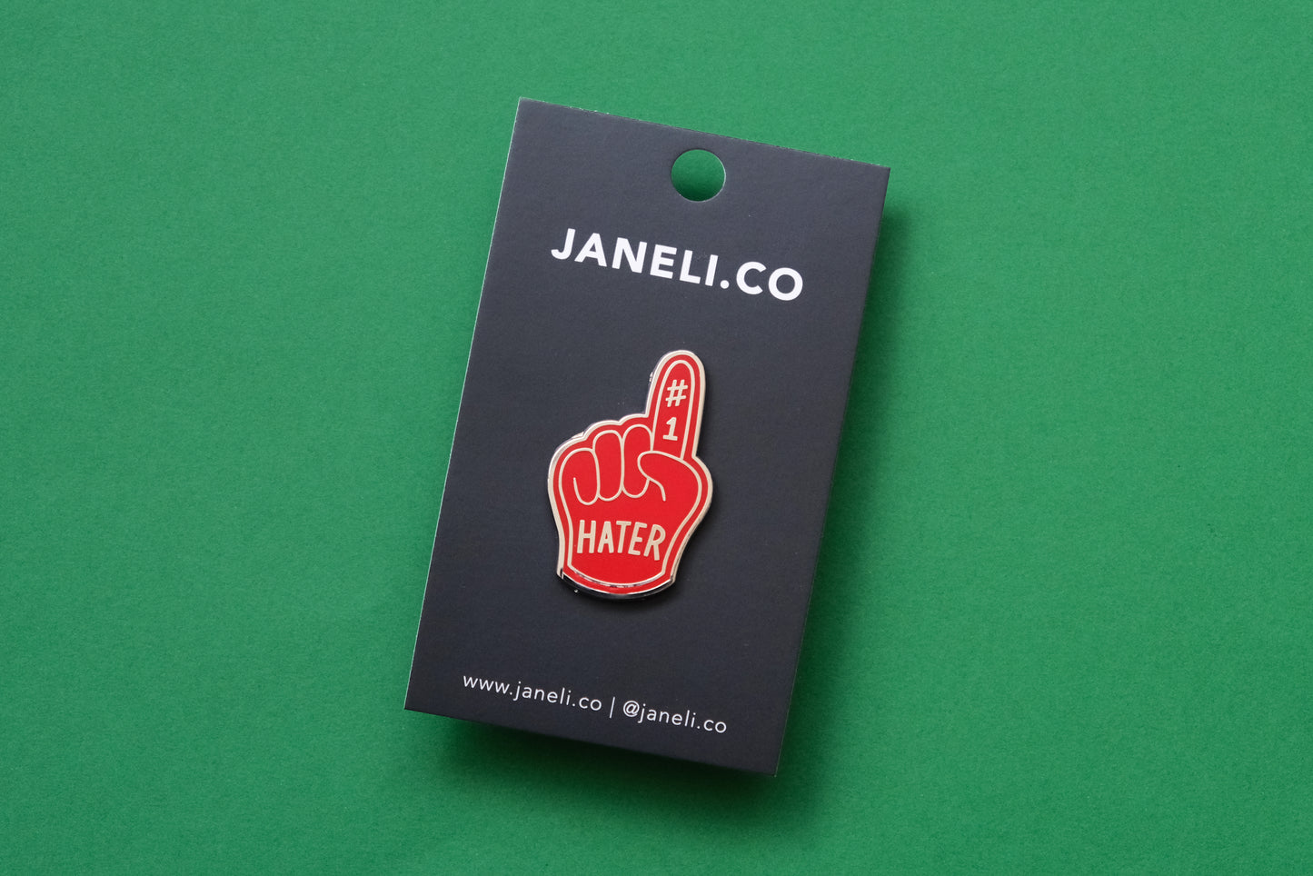 An enamel pin showing a rerd foam finger that says "#1 Hater" on a black JaneLi.Co backing card over a green background.