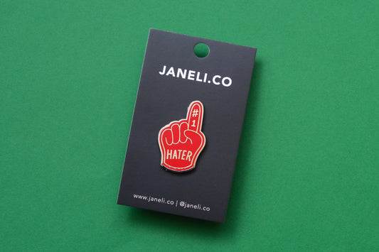 An enamel pin showing a rerd foam finger that says "#1 Hater" on a black JaneLi.Co backing card over a green background.