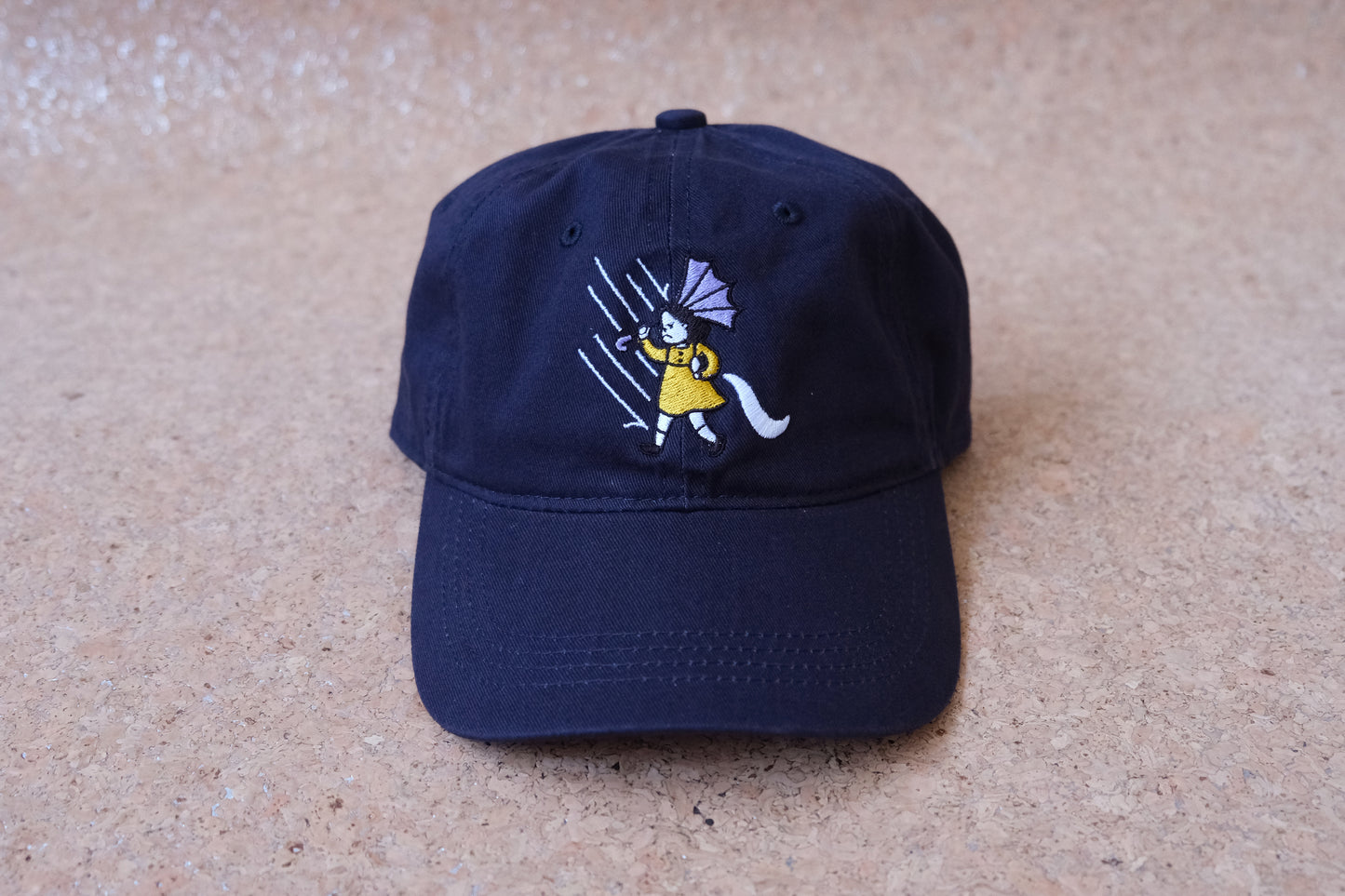 A photo of a navy dad hat with an embroidered grumpy salt girl over a tan background.