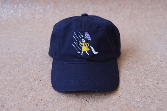 A photo of a navy dad hat with an embroidered grumpy salt girl over a tan background.