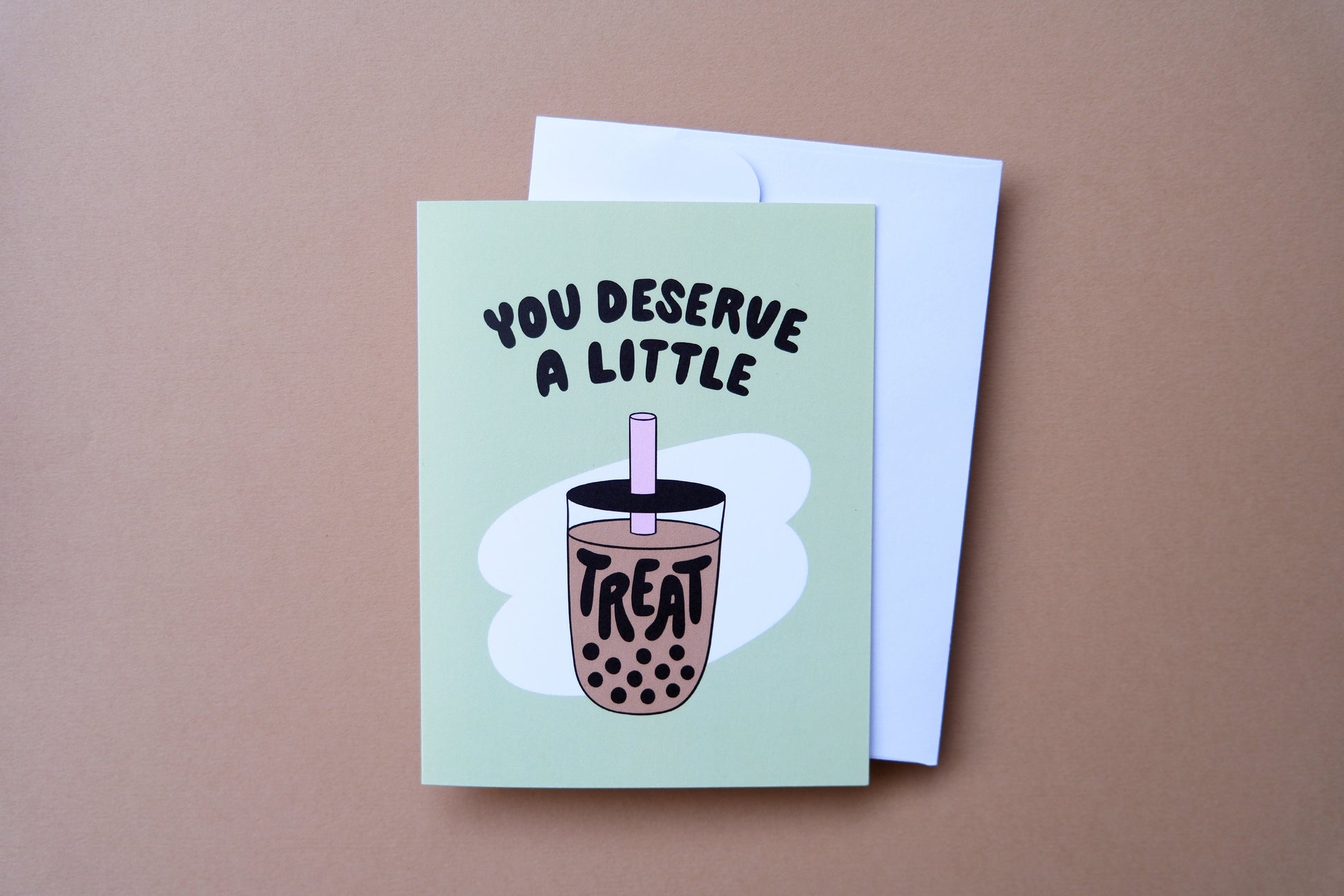 A photo of a green greeting card with a cup of boba that says "You deserve a little treat" and a white envelope on a tan background.