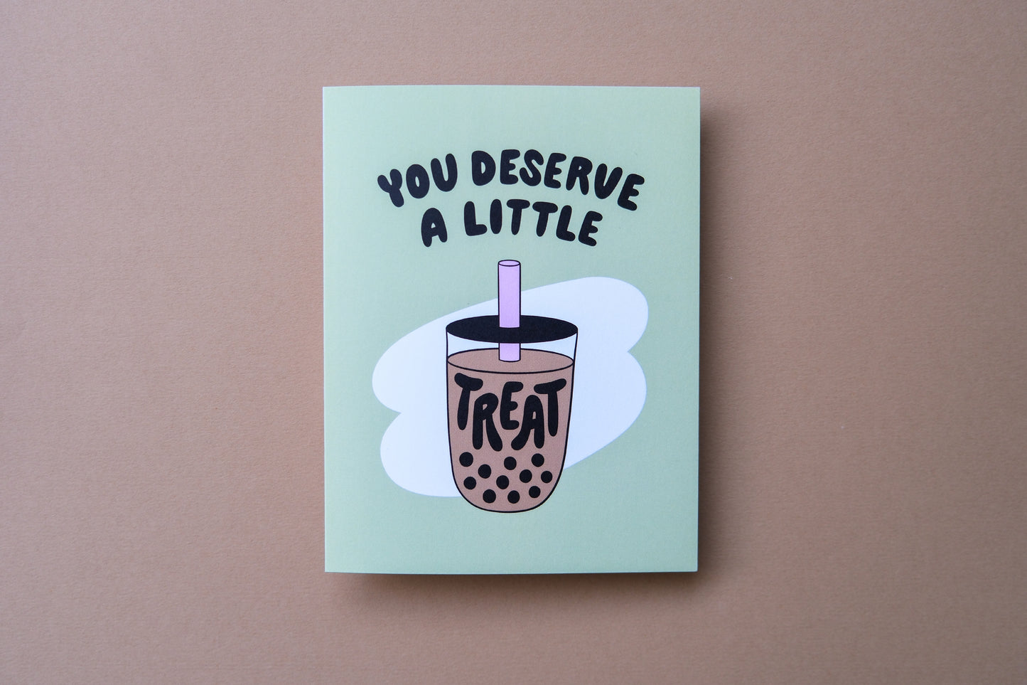 A photo of a green greeting card with a cup of boba that says "You deserve a little treat" on a tan background.