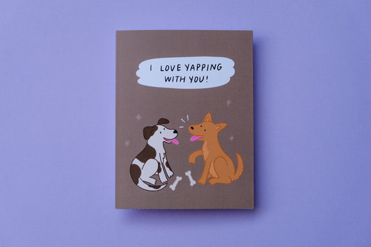 A photo of a chocolate greeting card that says "I love yapping with you!" with two pups chatting over a couple of bones on a purple background.