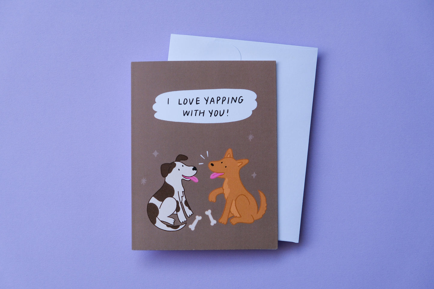 A photo of a chocolate greeting card that says "I love yapping with you!" with two pups chatting over a couple of bones and a white envelope on a purple background.