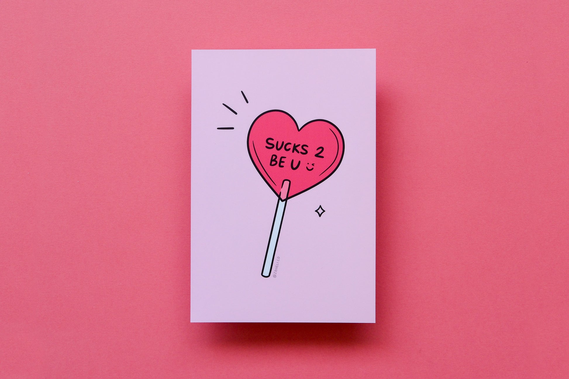 A JaneLi.Co mini print/postcard of a heart shaped lollipop that says "Sucks 2 Be U" over a pink background.