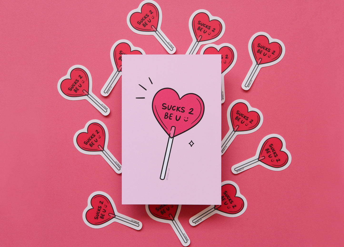 
A JaneLi.Co mini print/postcard of a heart shaped lollipop that says "Sucks 2 Be U" over scattered heart lollipop stickers on a pink background.