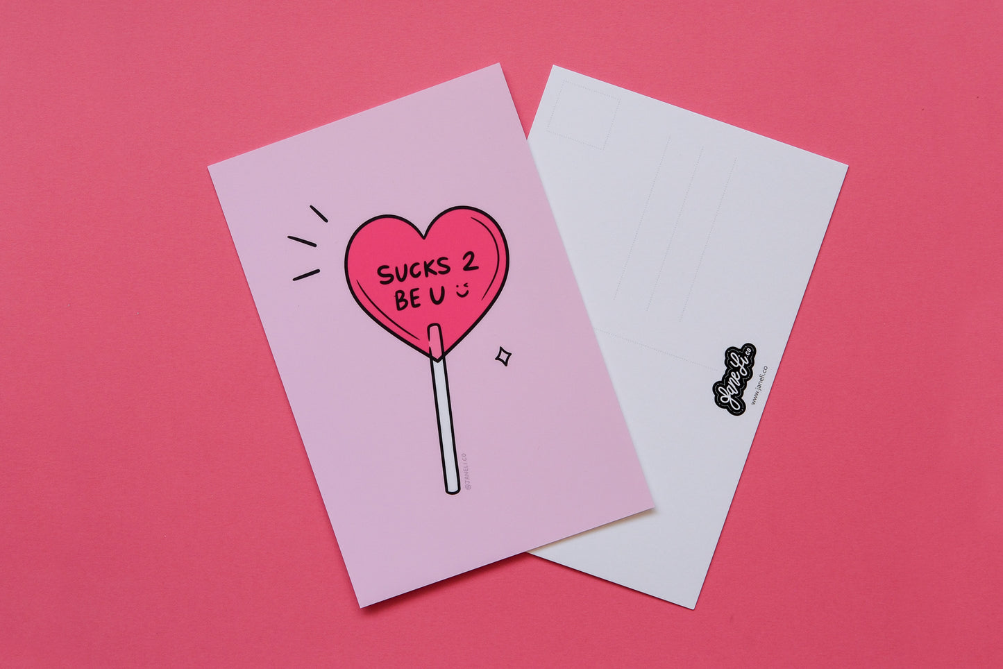 
A JaneLi.Co mini print/postcard of a heart shaped lollipop that says "Sucks 2 Be U" and a back postcard side of the same print over a pink background.