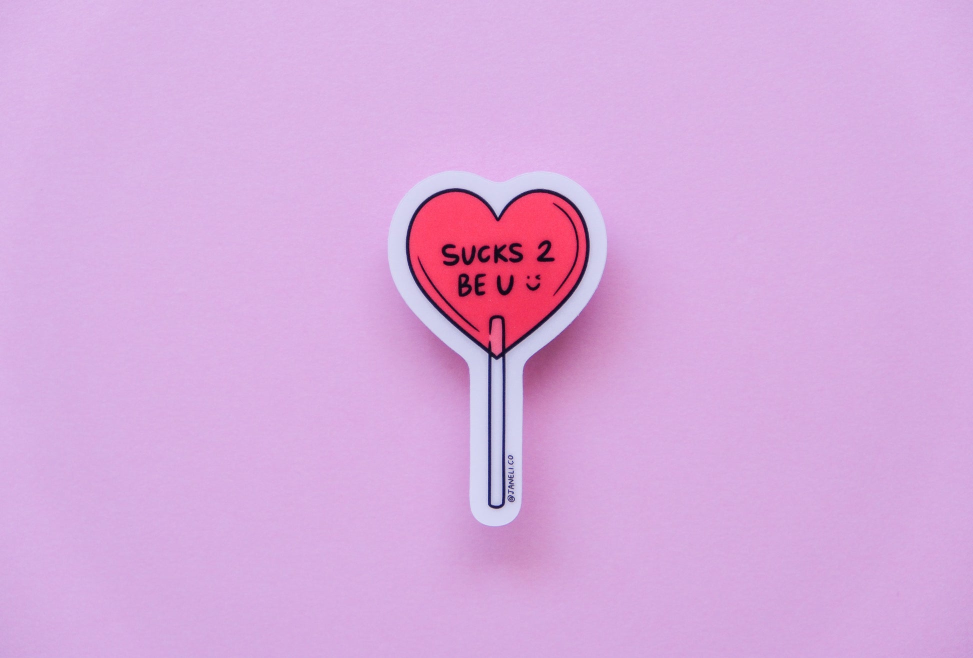 A JaneLi.Co sticker of a heart shaped lollipop that says "Sucks 2 Be U" over a pink background.