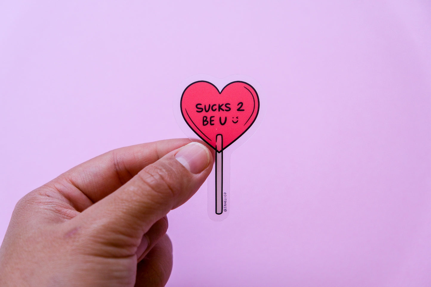 A hand holding a clear JaneLi.Co sticker of a heart shaped lollipop that says "Sucks 2 Be U" over a pink background.