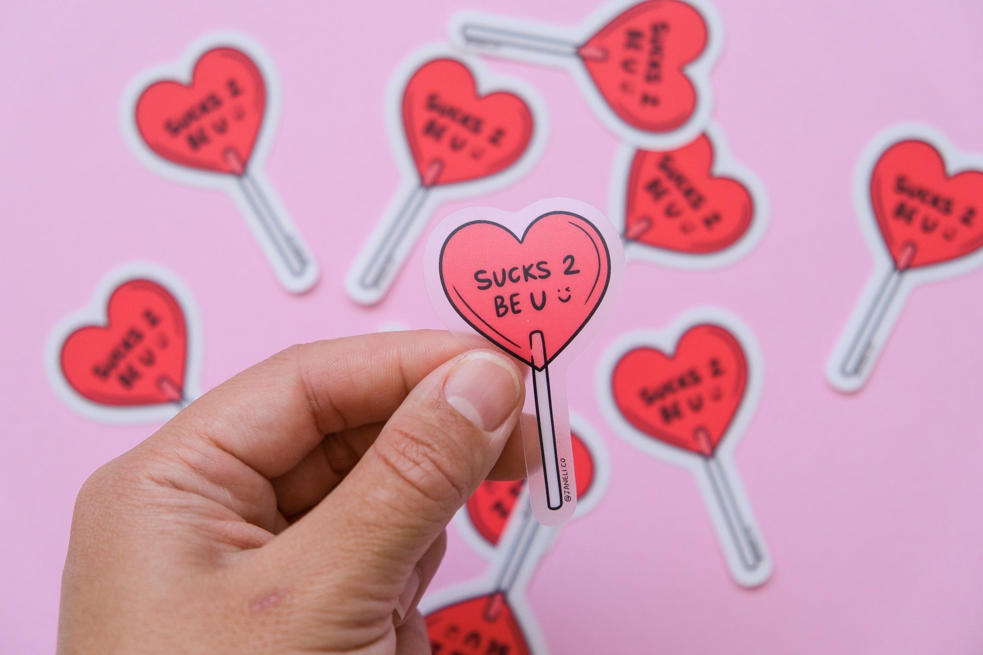 A hand holding a clear JaneLi.Co sticker of a heart shaped lollipop that says "Sucks 2 Be U" over a scattered pile of the same stickers on a  pink background.