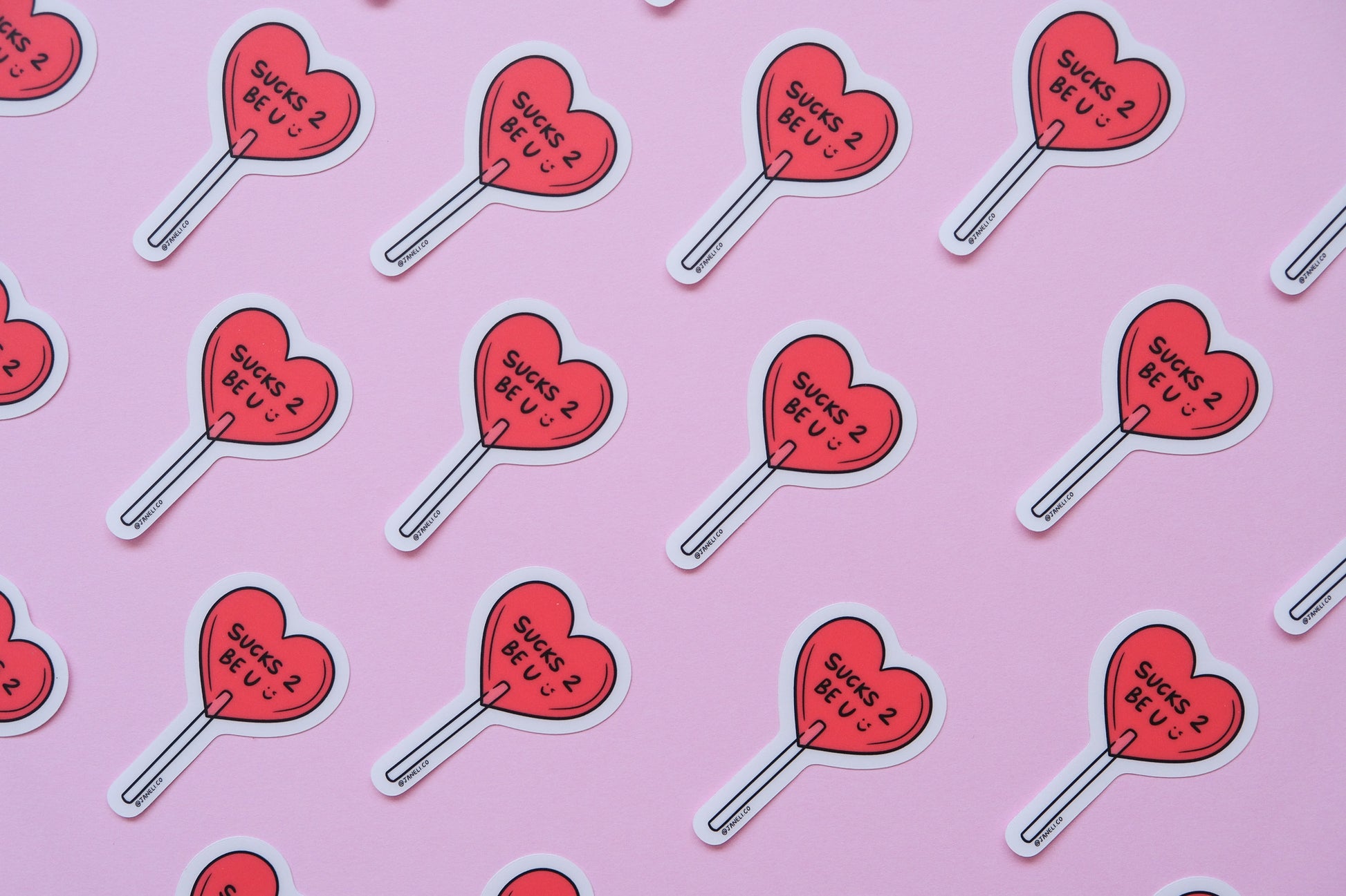 A grid of JaneLi.Co stickers of a heart shaped lollipops that say "Sucks 2 Be U" over a pink background.