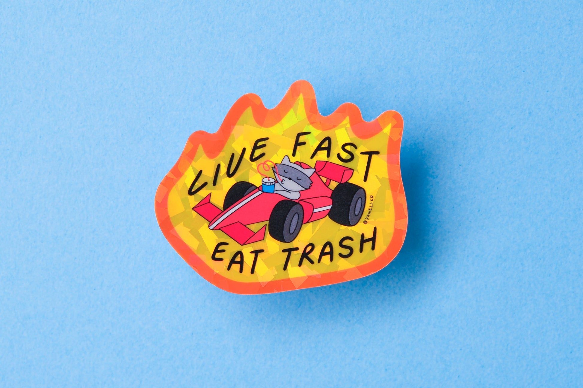 A JaneLi.Co sticker of a raccoon driving a racecar with flames while slurping a soda that says "Live Fast Eat Trash" over a blue background.