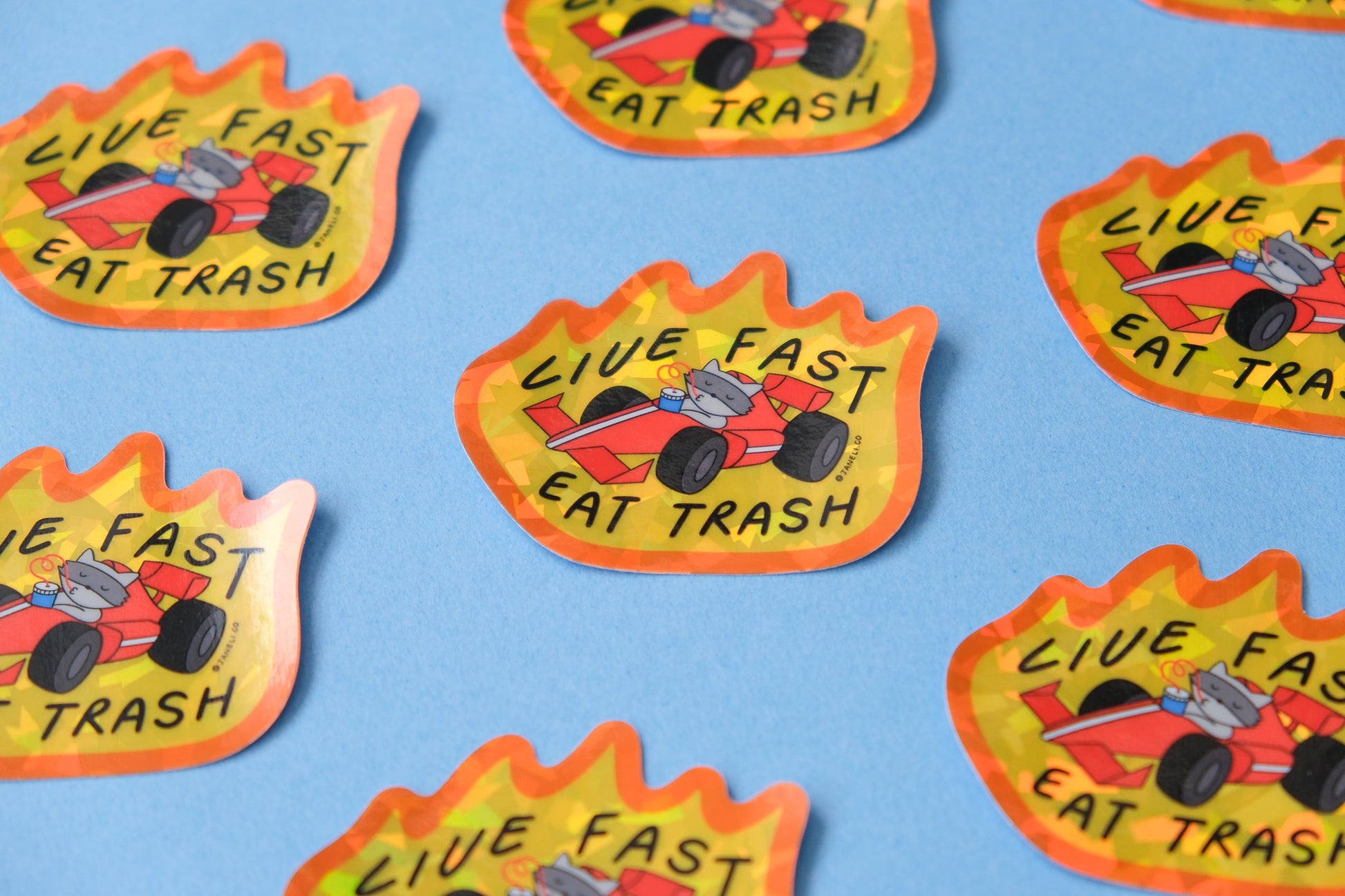 A grid of JaneLi.Co stickers of raccoons driving racecars with flames while slurping sodas that say "Live Fast Eat Trash" over a blue background.