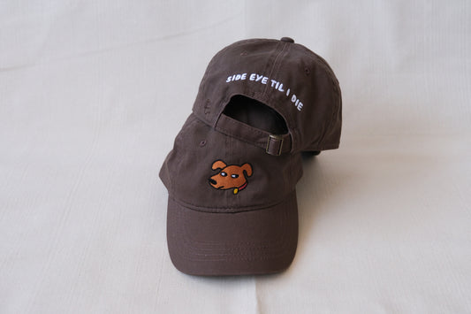 A photo of a chocolate brown dad hat with an embroidered dog giving side eye and the back of a second hat with "Side Eye Til I Die" embroidered on the back in white.