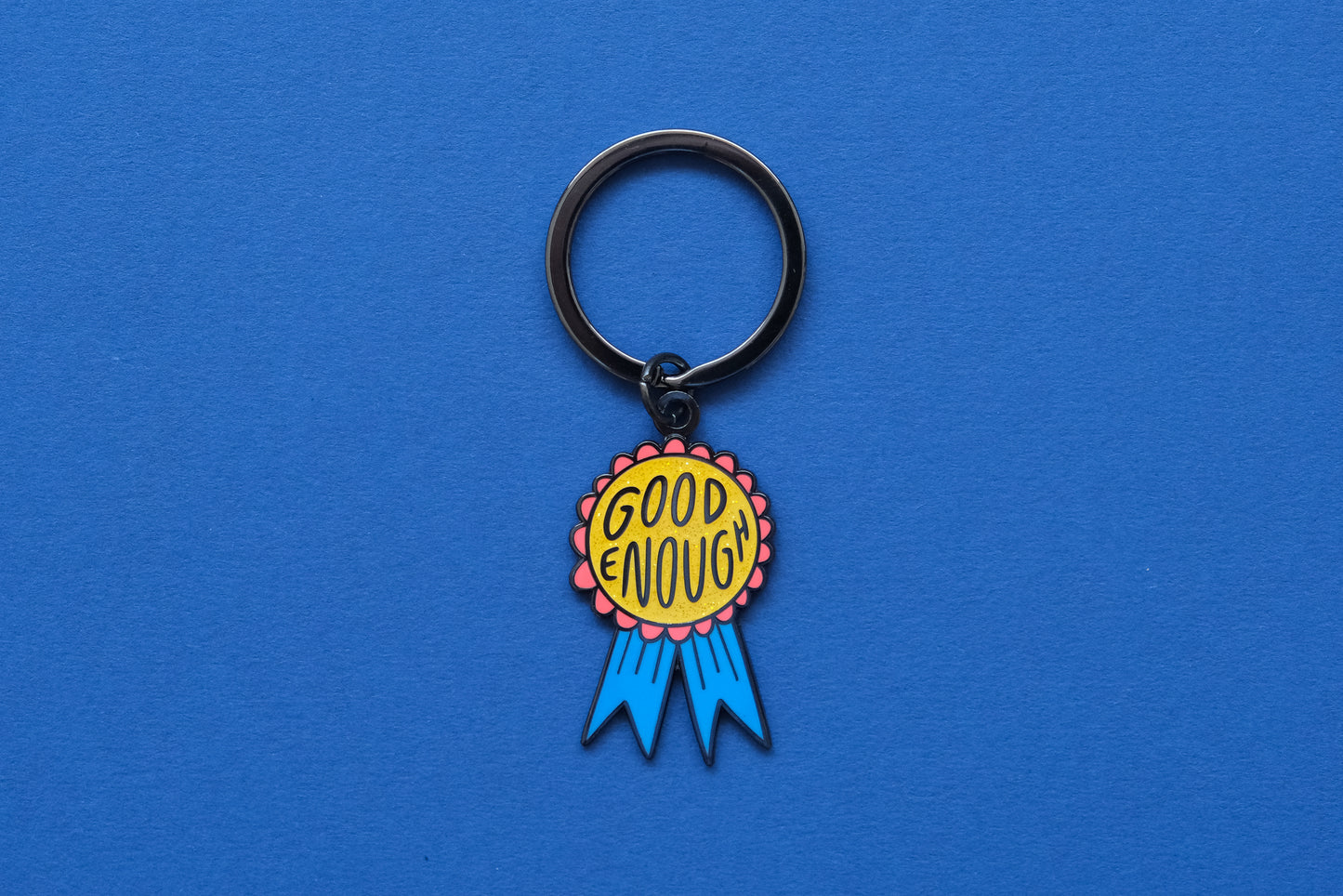 An enamel keychain showing a glittery red and blue award ribbon that says "Good Enough" over a blue background.