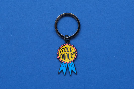 An enamel keychain showing a glittery red and blue award ribbon that says "Good Enough" over a blue background.