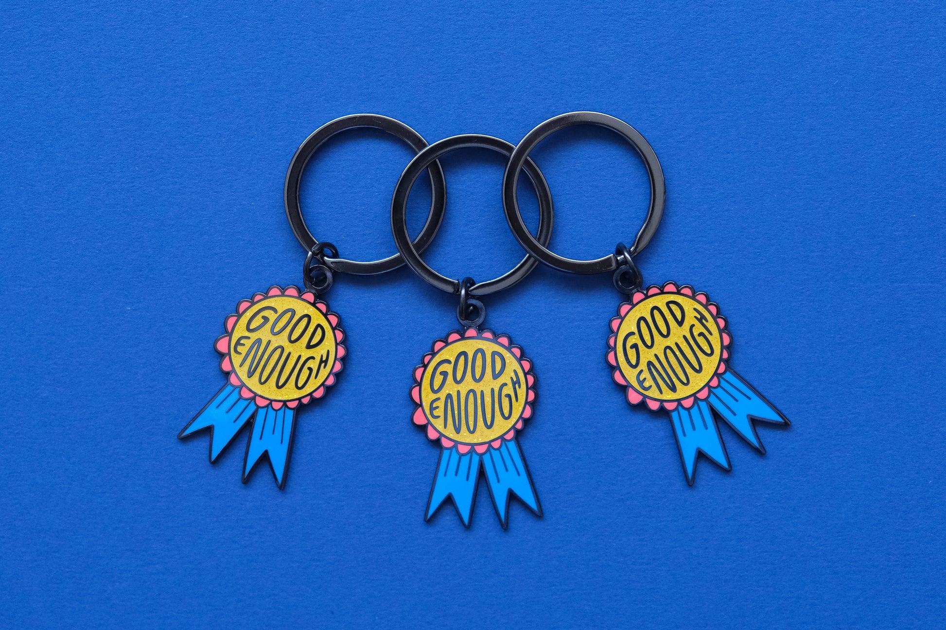 3 enamel keychains showing glittery red and blue award ribbons that says "Good Enough" over a blue background.