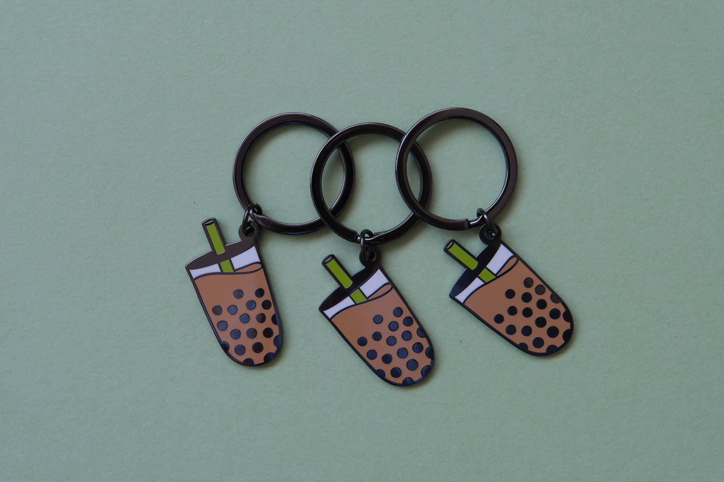 3 enamel keychains showing cups of boba with a  green straw over a green background.