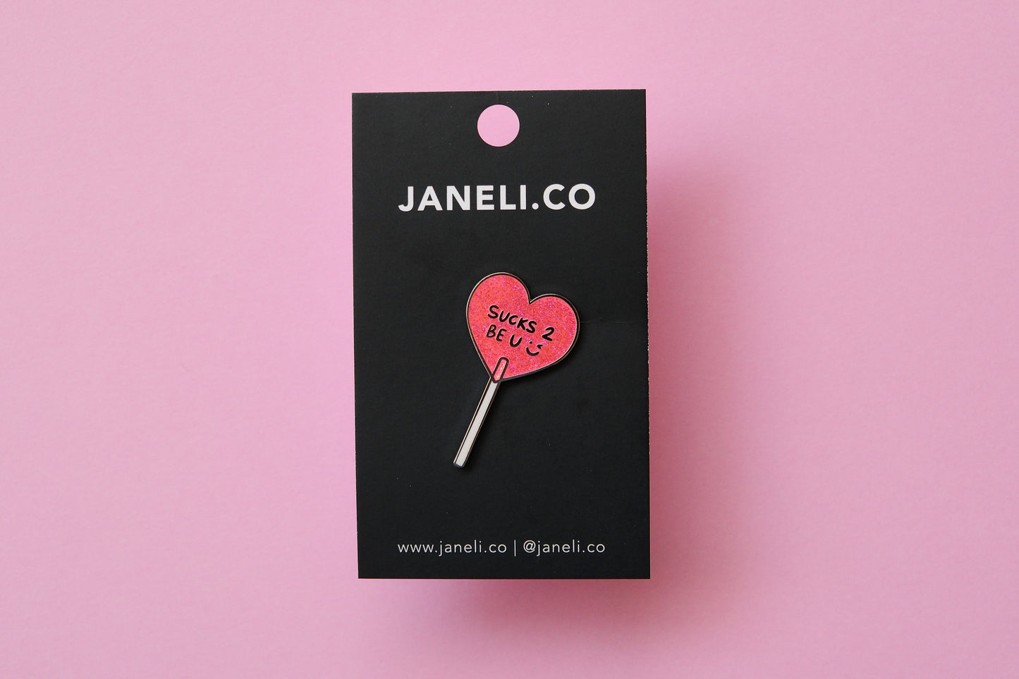 An enamel pin of a heart shaped lollipop that says "Sucks 2 Be U" on a black JaneLi.Co backing card over a pink background.