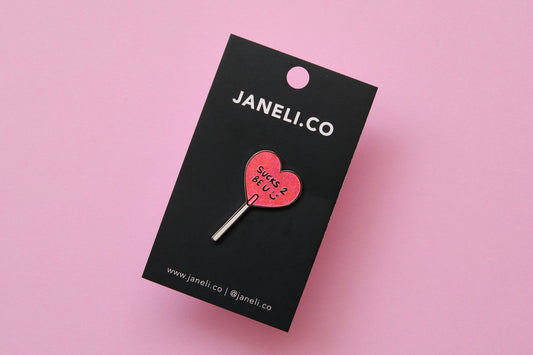 An enamel pin of a heart shaped lollipop that says "Sucks 2 Be U" on a black JaneLi.Co backing card over a pink background.