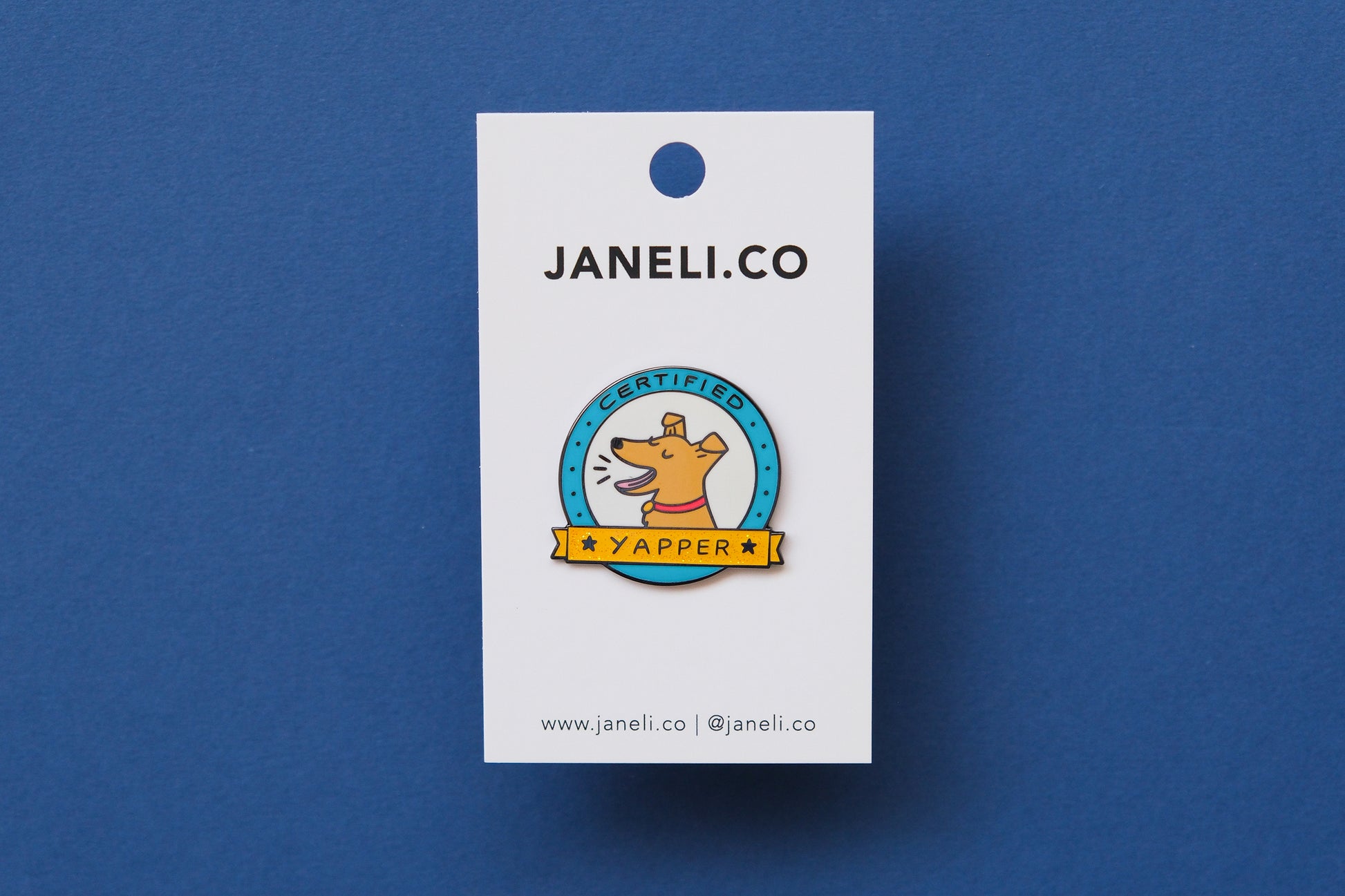 An enamel pin showing a barking dog that says "Certified Yapper" on a white JaneLi.Co backing card over a blue background.