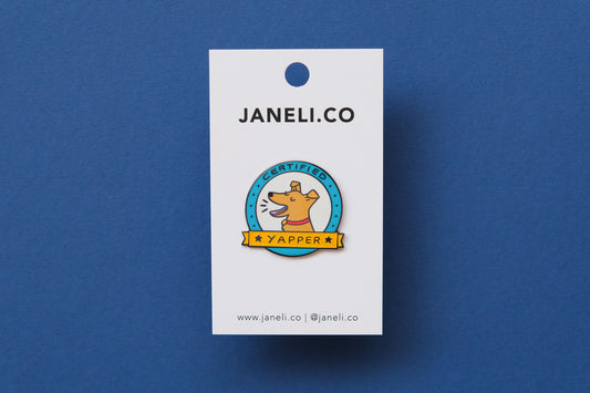 An enamel pin showing a barking dog that says "Certified Yapper" on a white JaneLi.Co backing card over a blue background.