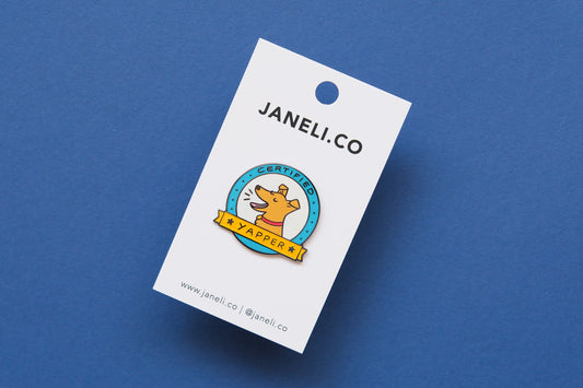 An enamel pin showing a barking dog that says "Certified Yapper" on a white JaneLi.Co backing card over a blue background.