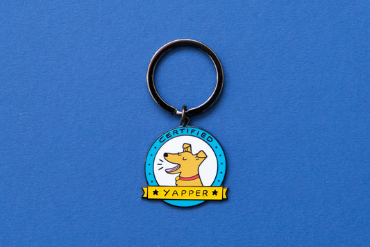 An enamel keychain showing a barking dog that says "Certified Yapper" over a blue background.