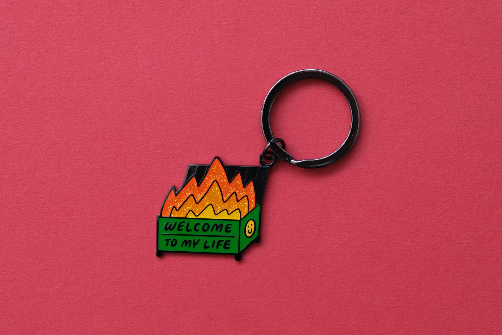 An enamel keychain showing a dumpster with glittery flames that says "Welcome to my life" over a red background.