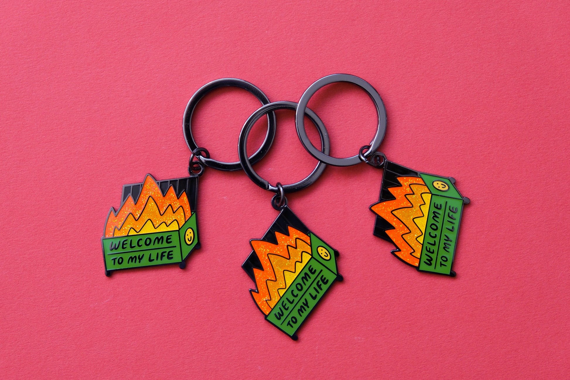 3 enamel keychains showing dumpsters with glittery flames that says "Welcome to my life" over a red background.