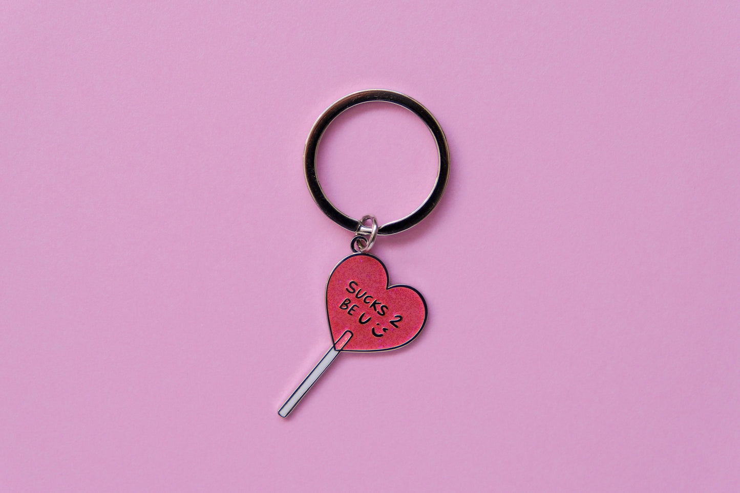 An enamel keychain of a heart shaped lollipop that says "Sucks 2 Be U" over a pink background.