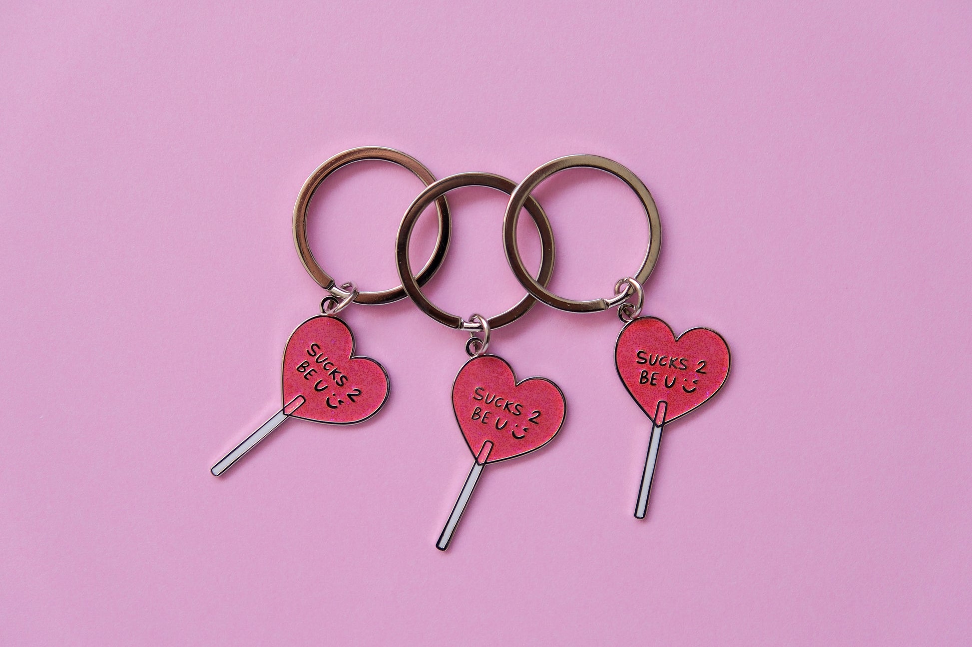 3 enamel keychains of heart shaped lollipops that say "Sucks 2 Be U" over a pink background.