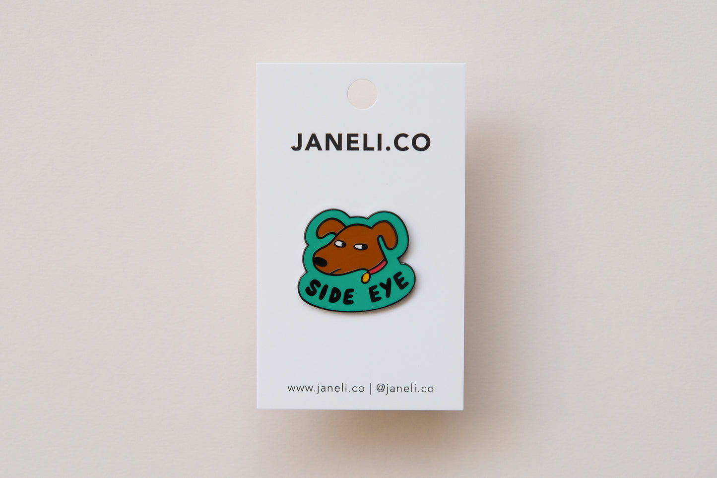 An enamel pin showing a sassy dog giving side eye that says "Side Eye" on a white JaneLi.Co backing card over a cream background.