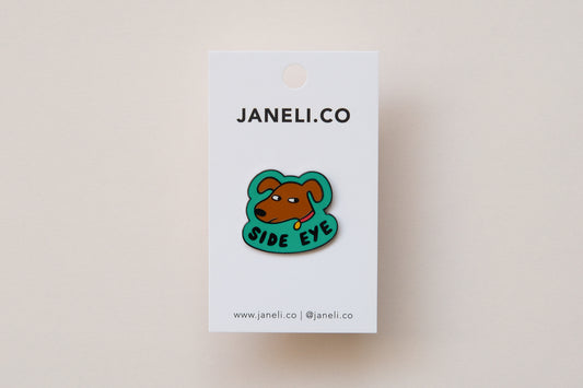 An enamel pin showing a sassy dog giving side eye that says "Side Eye" on a white JaneLi.Co backing card over a cream background.